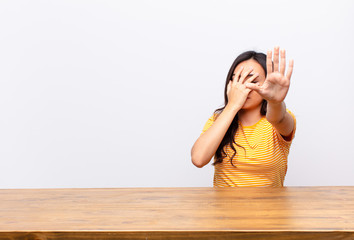 young latin pretty woman covering face with hand and putting other hand up front to stop camera,...