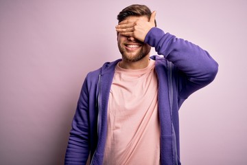 Young blond man with beard and blue eyes wearing purple sweatshirt over pink background smiling and laughing with hand on face covering eyes for surprise. Blind concept.