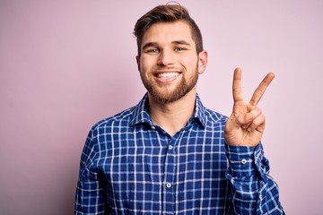 Fototapeta premium Young handsome blond man with beard and blue eyes wearing casual shirt standing smiling with happy face winking at the camera doing victory sign. Number two.