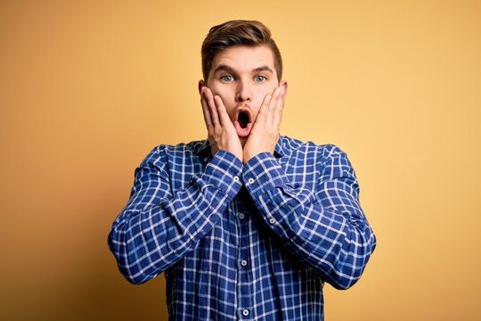 Young blond businessman with beard and blue eyes wearing shirt over yellow background afraid and shocked, surprise and amazed expression with hands on face