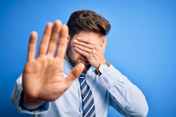 Young blond businessman with beard and blue eyes wearing elegant shirt and tie standing covering eyes with hands and doing stop gesture with sad and fear expression. Embarrassed and negative concept.