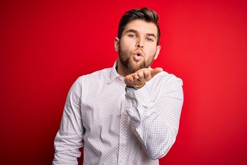 Young blond businessman with beard and blue eyes wearing elegant shirt over red background looking at the camera blowing a kiss with hand on air being lovely and sexy. Love expression.