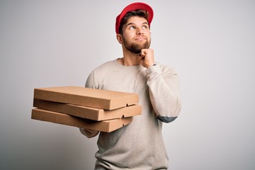 Young blond delivery man with beard and blue eyes holding cardboards of pizza serious face thinking...