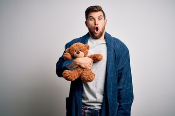 Young blond man with beard and blue eyes wearing pajama holding teddy bear scared in shock with a...