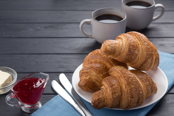 Fresh croissants on a black wooden table. Served with coffee, coffeepot, butter, jam, blue napkin. Fresh French Baked Croissants. Warm Fresh Buttery Rolls. Free space for text. 