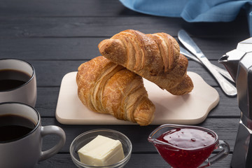 Fresh croissants on a black wooden table. Served with coffee, coffeepot, butter, jam, blue napkin. Fresh French Baked Croissants. Warm Fresh Buttery Rolls. Free space for text. 