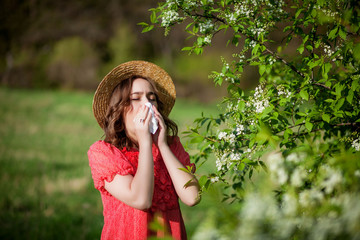 Young girl blowing nose and sneezing in tissue in front of blooming tree. Seasonal allergens...