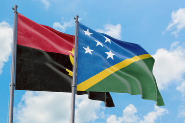 Solomon Islands and Angola flags waving in the wind against white cloudy blue sky together....