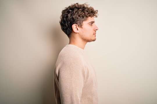 Young blond handsome man with curly hair wearing casual sweater over white background looking to side, relax profile pose with natural face with confident smile.