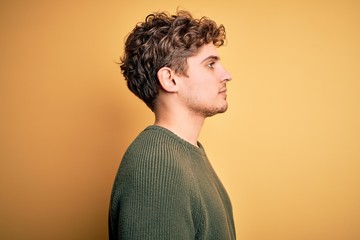 Young blond handsome man with curly hair wearing green sweater over yellow background looking to side, relax profile pose with natural face with confident smile.