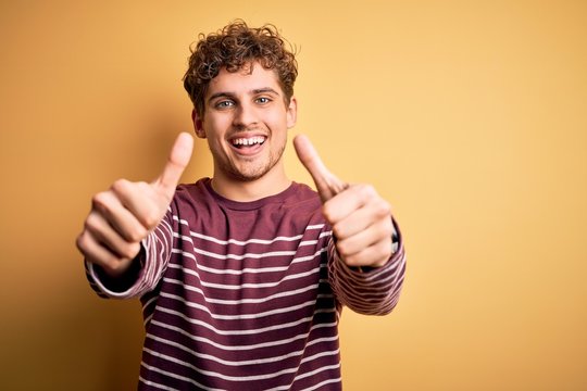 Young blond handsome man with curly hair wearing casual striped sweater approving doing positive gesture with hand, thumbs up smiling and happy for success. Winner gesture.