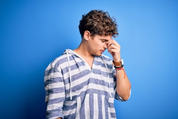 Young blond handsome man with curly hair wearing casual striped sweatshirt tired rubbing nose and eyes feeling fatigue and headache. Stress and frustration concept.