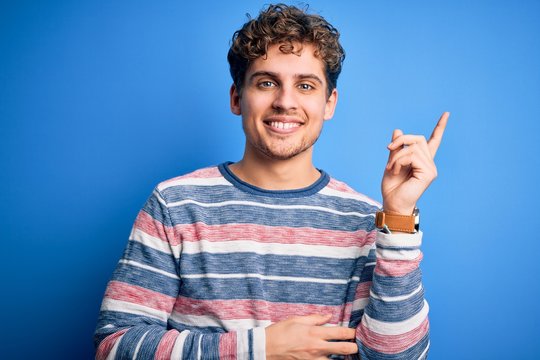 Young blond handsome man with curly hair wearing striped sweater over blue background with a big smile on face, pointing with hand and finger to the side looking at the camera.