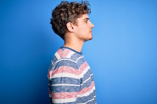 Young blond handsome man with curly hair wearing striped sweater over blue background looking to side, relax profile pose with natural face with confident smile.