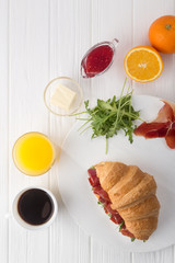 Fresh croissant sandwich with ham, cheese and arugula on white wooden table. Served with Coffee, cheese, ham, orange juice, jam. Fresh French Baked Croissants. Free space for text. View from above.