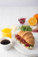 Fresh croissant sandwich with ham, cheese and arugula on white wooden table. Served with Coffee, cheese, ham, orange juice, jam. Fresh French Baked Croissants. Free space for text. 