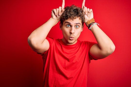 Young blond handsome man with curly hair wearing casual t-shirt over red background doing funny gesture with finger over head as bull horns