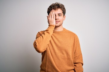 Young blond handsome man with curly hair wearing casual sweater over white background covering one eye with hand, confident smile on face and surprise emotion.