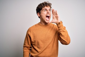 Young blond handsome man with curly hair wearing casual sweater over white background shouting and screaming loud to side with hand on mouth. Communication concept.