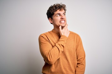 Young blond handsome man with curly hair wearing casual sweater over white background Thinking worried about a question, concerned and nervous with hand on chin