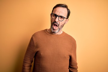 Middle age hoary man wearing brown sweater and glasses over isolated yellow background In shock face, looking skeptical and sarcastic, surprised with open mouth