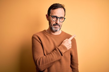 Fototapeta premium Middle age hoary man wearing brown sweater and glasses over isolated yellow background Pointing with hand finger to the side showing advertisement, serious and calm face