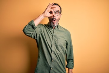 Middle age hoary man wearing casual green shirt and glasses over isolated yellow background doing ok gesture shocked with surprised face, eye looking through fingers. Unbelieving expression.