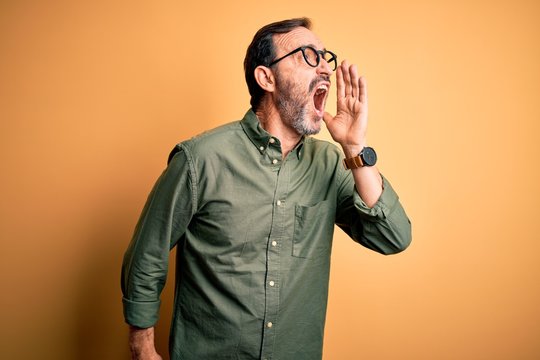 Middle age hoary man wearing casual green shirt and glasses over isolated yellow background shouting and screaming loud to side with hand on mouth. Communication concept.