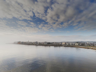 Aerial drone view on Salthill from Galway bay, Cloudy blue sky, Calm ocean water. Light fog on left side.