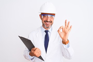 Senior engineer man wearing helmet glasses holding clipboard over isolated white background doing ok sign with fingers, excellent symbol