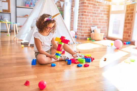 Beautiful toddler wearing glasses and unicorn diadem sitting playing with building blocks at kindergarten