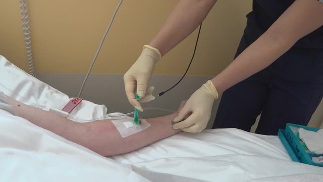 The nurse's hands in sterile gloves are injected into the venous catheter. You can see the patient's hand with a catheter and a drip tube and two nurse's hands, in the background usb-C connector.