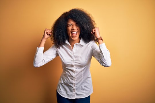 Young beautiful african american elegant woman with afro hair standing over yellow background angry and mad raising fists frustrated and furious while shouting with anger. Rage and aggressive concept.