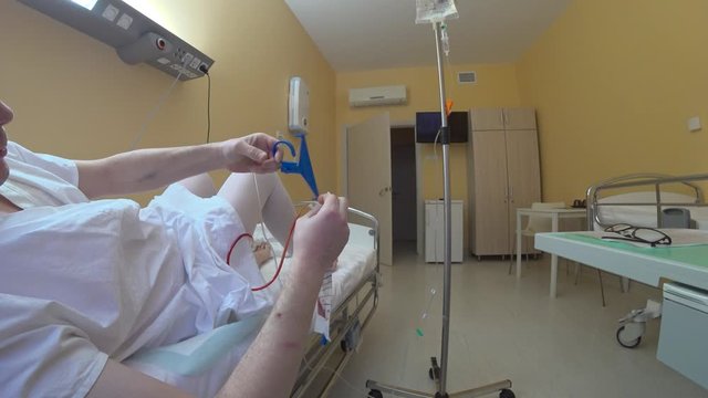 Patient corrects drainage system after surgery. Ward in the hospital, the patient after surgery with catheters and tubes in the body with difficulty independently performs simple actions.