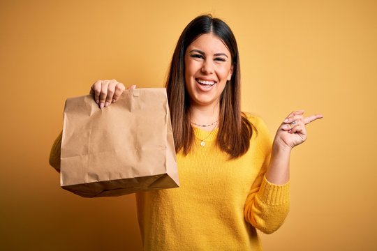 Young Beautiful Woman Holding Take Away Paper Bag From Delivery Over Yellow Background Very Happy Pointing With Hand And Finger To The Side