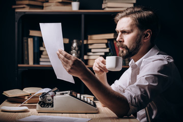 Serious pensive penman sitting at desk, drinking coffee and reading his recent text