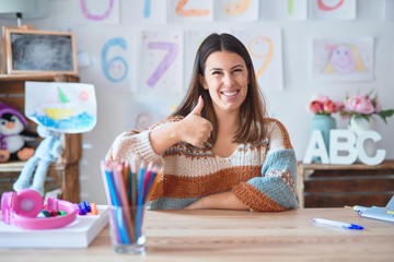 Young beautiful teacher woman wearing sweater and glasses sitting on desk at kindergarten doing...