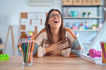 Young beautiful teacher woman wearing sweater and glasses sitting on desk at kindergarten smiling...