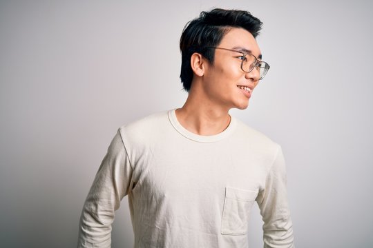 Young Handsome Chinese Man Wearing Casual T-shirt And Glasses Over White Background Looking Away To Side With Smile On Face, Natural Expression. Laughing Confident.