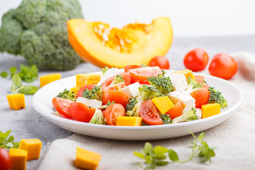 Vegetarian salad with broccoli, tomatoes, feta cheese, and pumpkin on white ceramic plate on a gray concrete background, side view, close up.