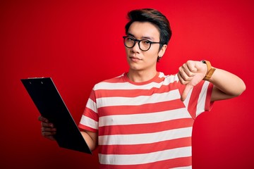 Young handsome chinese inspector man wearing glasses holding clipboard checklist with angry face,...