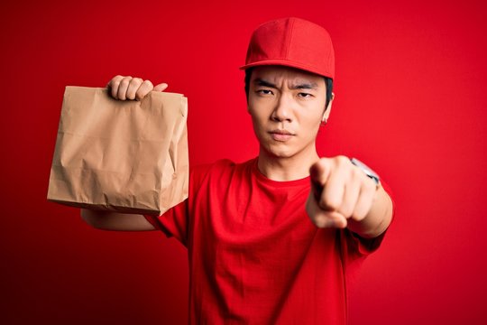 Young Handsome Chinese Delivery Man Holding Takeaway Paper Bag With Food Pointing With Finger To The Camera And To You, Hand Sign, Positive And Confident Gesture From The Front