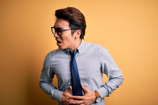 Young handsome chinese businessman wearing glasses and tie over yellow background with hand on stomach because nausea, painful disease feeling unwell. Ache concept.