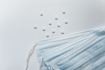 scattered small white round pill pills with a blue medical mask next to a white even background