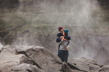 Young stylish couple in love kissing together near famous Icelandic landscape Dettifoss waterfall. Traditional wool sweaters, hat, red hair, gray skirt. Dramatic landscape, cold weather in Iceland.