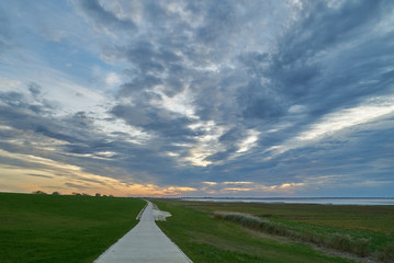 rural lane next to a dike at in the community Butjadingen, district Wesermarsch (Germany) under scenic sunset sky with mystic grey clouds