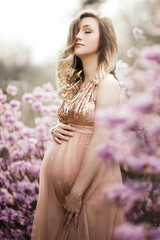 a delicate curly-haired pregnant blonde in full bloom of purple Rhododendron flowers in a peach tulle glittering dress hugs her big tummy and smiles, spring pregnancy