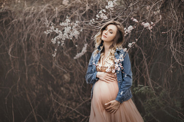 Fototapeta premium a delicate curly-haired pregnant blonde in full bloom of sakura flowers in a peach tulle glittering dress hugs her big tummy and smiles with closed eyes, spring pregnancy