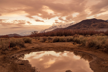 Fototapeta na wymiar early morning clouds, hills, snowy mountains reflected in rain puddle in dirt road