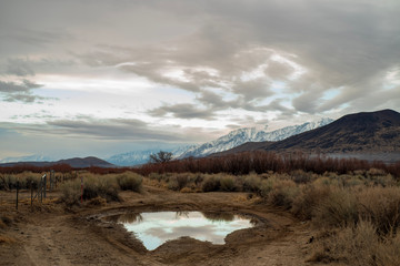 early morning clouds, hills, snowy mountains reflected in rain puddle in dirt road